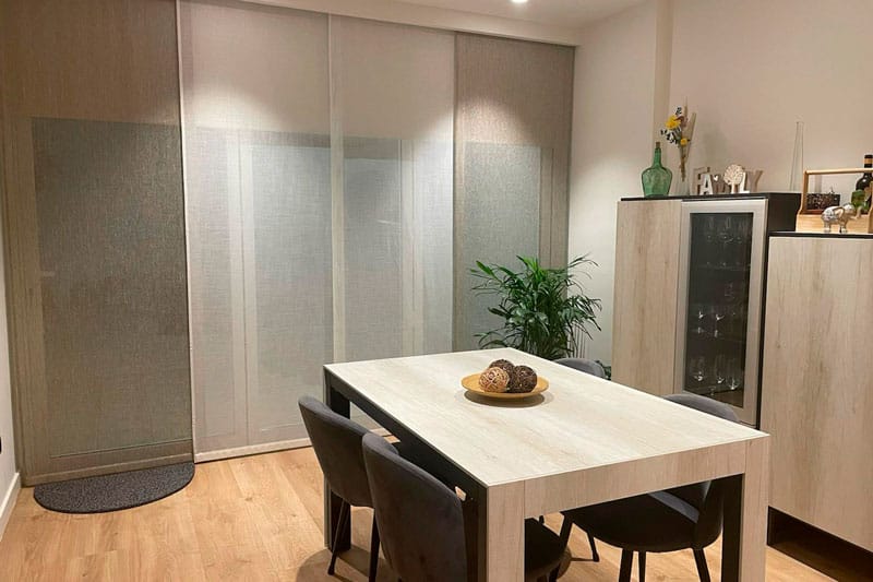 Comedor con mesa de madera clara, sillas negras y cortinas translúcidas en vivienda reformada en Chinchón.